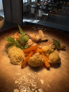 A plated dish featuring mashed potatoes, grilled meat topped with creamy sauce, sautéed mushrooms, roasted carrots, and cauliflower florets, garnished with fresh greens