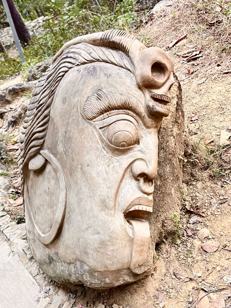 A large, carved stone sculpture of a face with exaggerated features, lying on a sandy and rocky surface outdoors. The sculpture has prominent eyebrows, a wide open mouth, and detailed hair, giving it a dramatic and expressive appearance