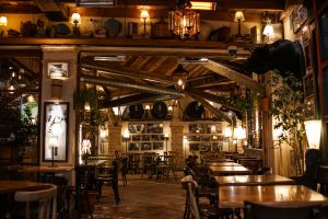 A rustic, warmly lit interior of a restaurant with wooden beams, tables, and chairs. The space features vintage decor, plants, wall art, and framed photographs. A mounted bull's head and various knick-knacks are displayed on upper shelves.