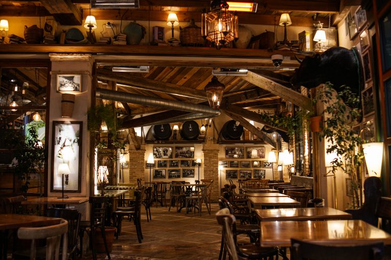 A rustic, warmly lit interior of a restaurant with wooden beams, tables, and chairs. The space features vintage decor, plants, wall art, and framed photographs. A mounted bull’s head and various knick-knacks are displayed on upper shelves.