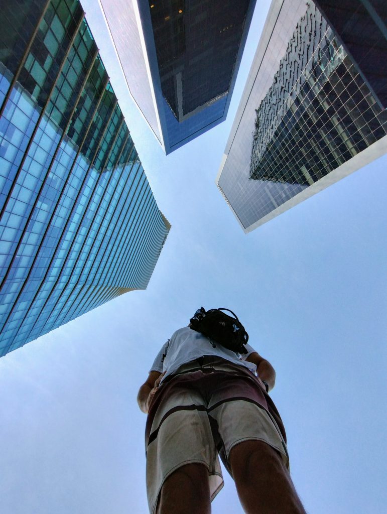 A low-angle view of a person wearing a backpack, looking up at several tall skyscrapers with glass exteriors against a clear blue sky.