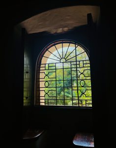 A wooden arched window with intricate ironwork patterns that overlooks lush green foliage outside. The interior is dimly lit, emphasizing the contrast with the bright outdoor scenery.