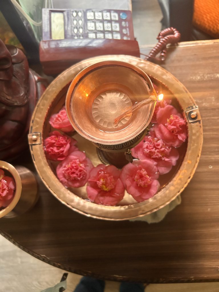 A copper bowl filled with water and pink flowers, with a small oil lamp floating in the center, surrounded by a decorative copper container on a wooden surface. In the background is a device with buttons.