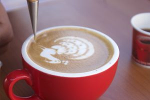 A red cup of coffee on a table with intricate latte art depicting a peacock, created using milk foam, with a metal art tool forming details.