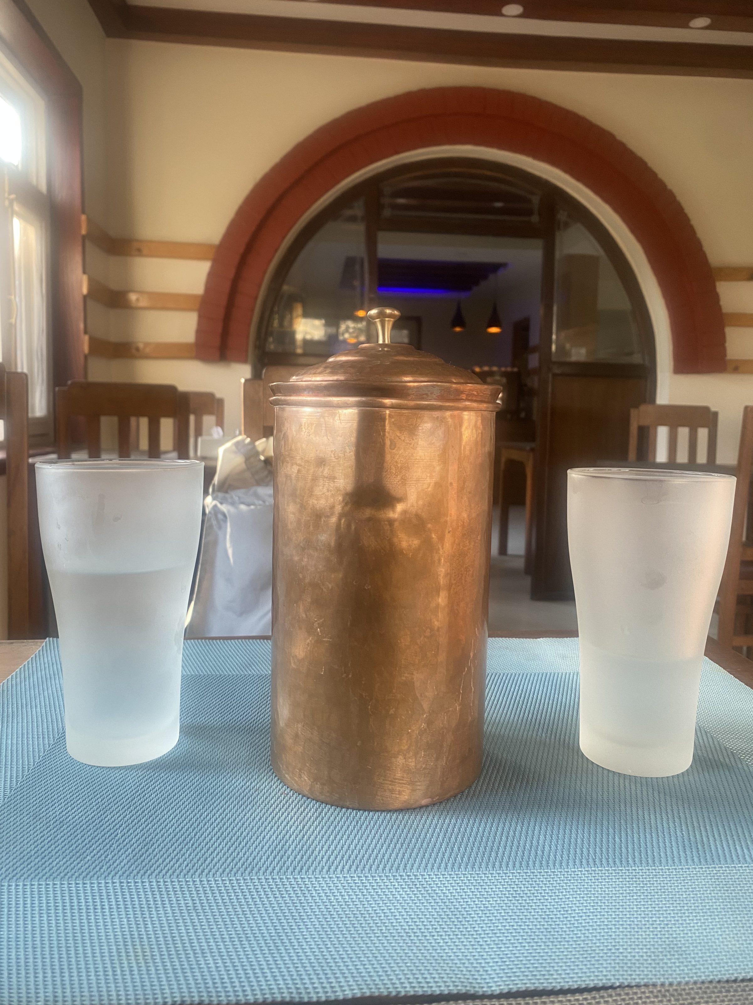 A copper container with a lid is placed on a blue mat, flanked by two frosted glass tumblers filled with water. The setting is indoors, featuring wooden chairs and an archway in the background.