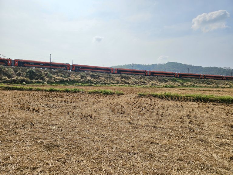 A long, orange intercity train travels across a rural landscape with harvested fields under a partly cloudy sky.