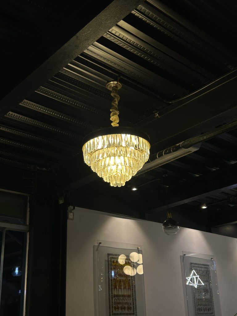 A large, ornate chandelier hangs from a dark, metal ceiling illuminating a room with framed artwork on the walls.