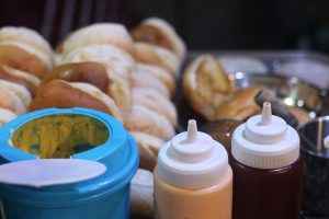 A close-up of various sauces in squeeze bottles and an open container of mustard in front of a stack of hamburger buns.