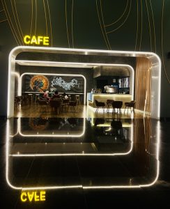 A modern, stylish cafe interior with glowing LED lights outlining the doorway and ceiling. The word "CAFE" is illuminated in yellow above the entrance. Inside, there are tables and chairs with a mural on the far wall. 