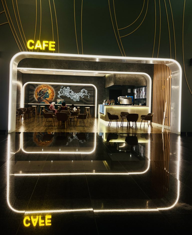 A modern, stylish cafe interior with glowing LED lights outlining the doorway and ceiling. The word “CAFE” is illuminated in yellow above the entrance. Inside, there are tables and chairs with a mural on the far wall.