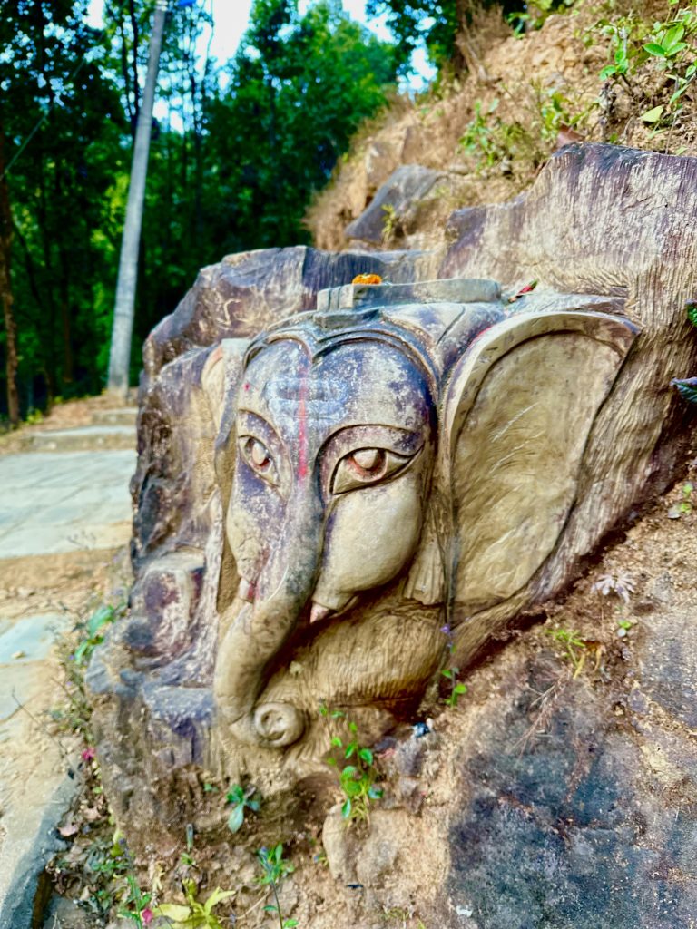 A carved Lord Ganesha elephant head sculpture is embedded in a rocky hillside surrounded by greenery, with trees visible in the background.