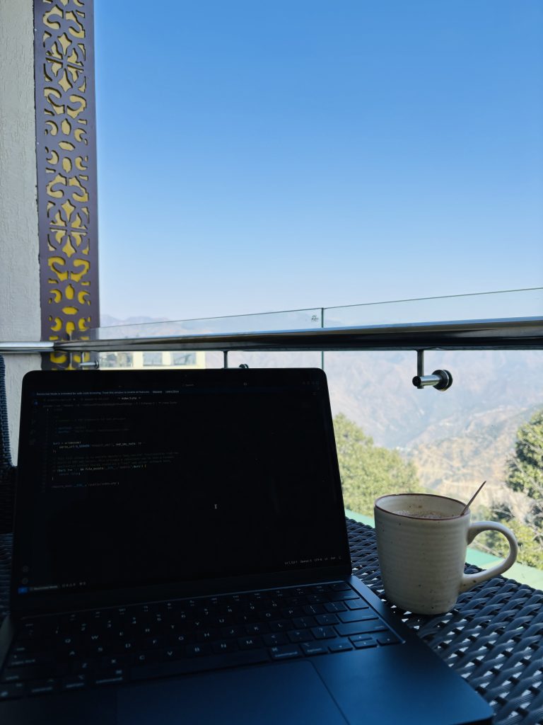 A laptop displaying code is placed on a woven outdoor table, with a mug of coffee beside it. The background features a clear blue sky and mountain landscape visible from a balcony with decorative railing elements.