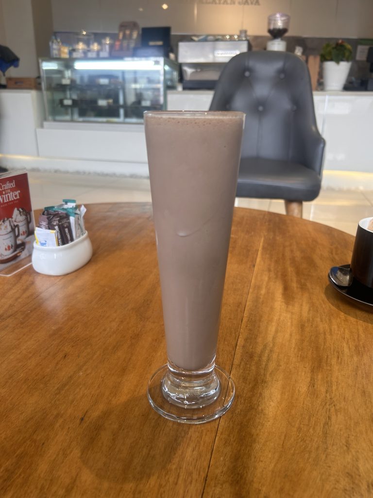 A tall glass of chocolate milkshake on a wooden table in a cozy cafe setting. In the background is a gray upholstered chair and a counter with a display case and coffee-making equipment.