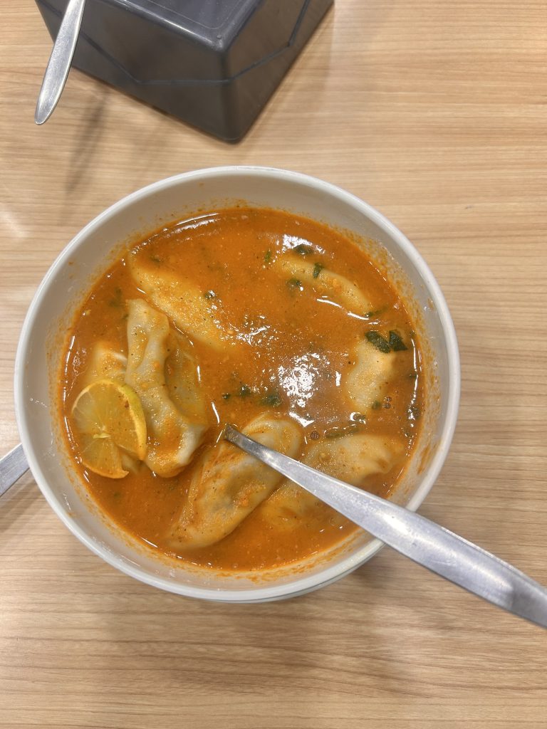 A bowl of bright orange soup with dumplings and sliced lemon, garnished with herbs, and served with spoons on a wooden table.