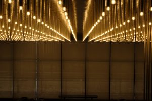 A dark, enclosed space with a grid of exposed light bulbs on the ceiling, creating an illuminated pathway effect with reflections on the walls and floor.