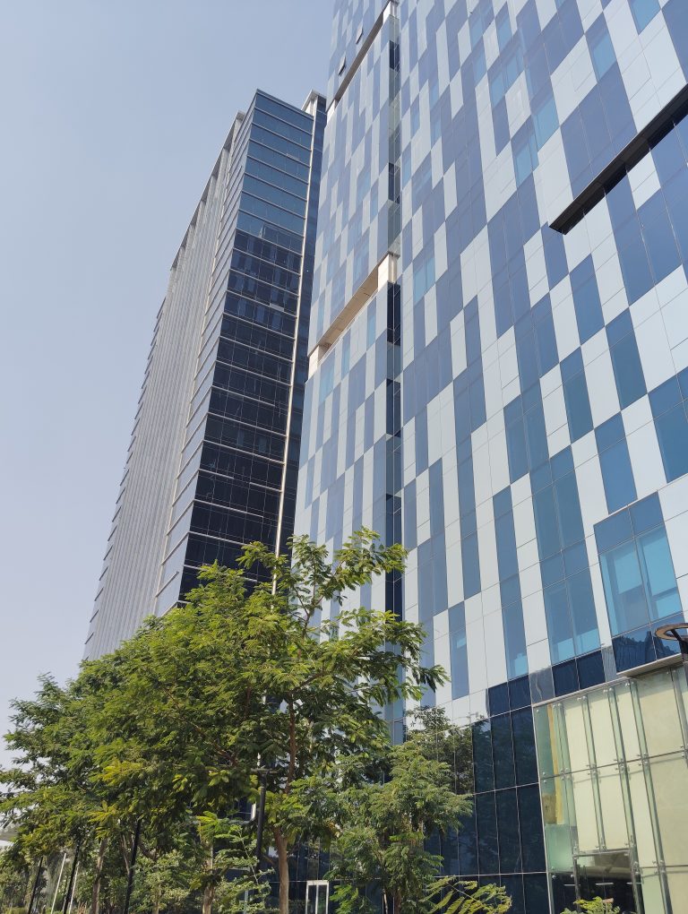 Tall modern buildings with reflective glass facades, surrounded by green trees under a clear blue sky.