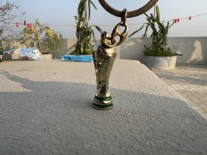 A small gold-colored FIFA World Cup trophy keychain sitting on a concrete surface, with a clothesline and potted plants visible in the blurred background.
