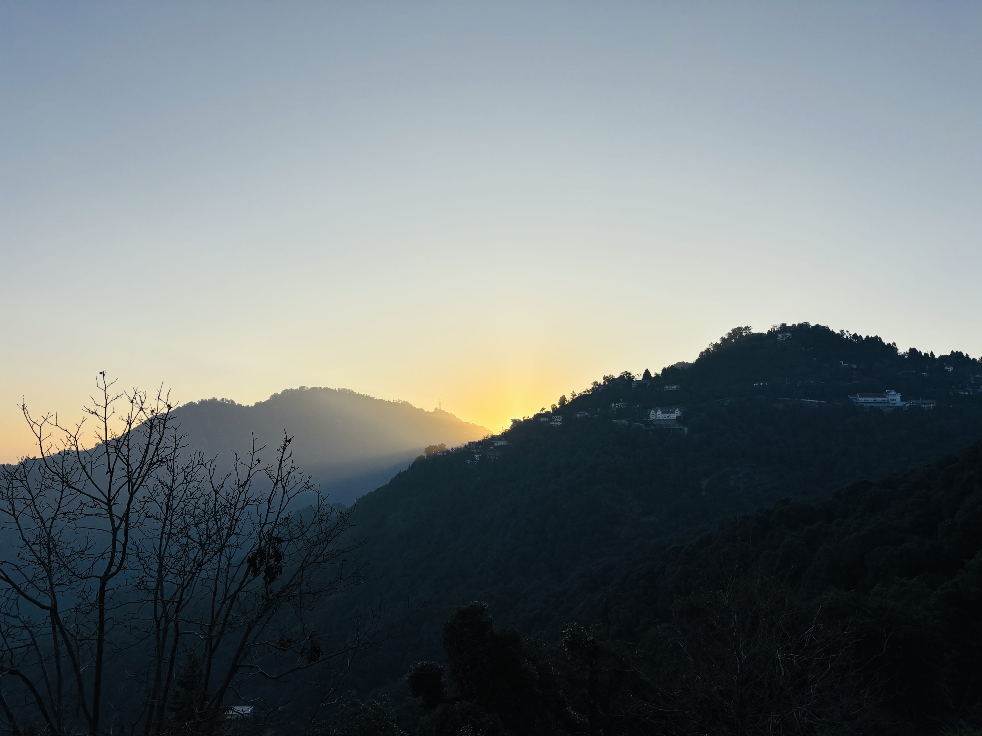 A scenic view of a sunrise behind dark silhouettes of hills, with a few trees and buildings visible against a gradient sky transitioning from a light orange to pale blue.