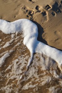 Waves with white foam washing up on a sandy beach, with footprints visible in the sand.