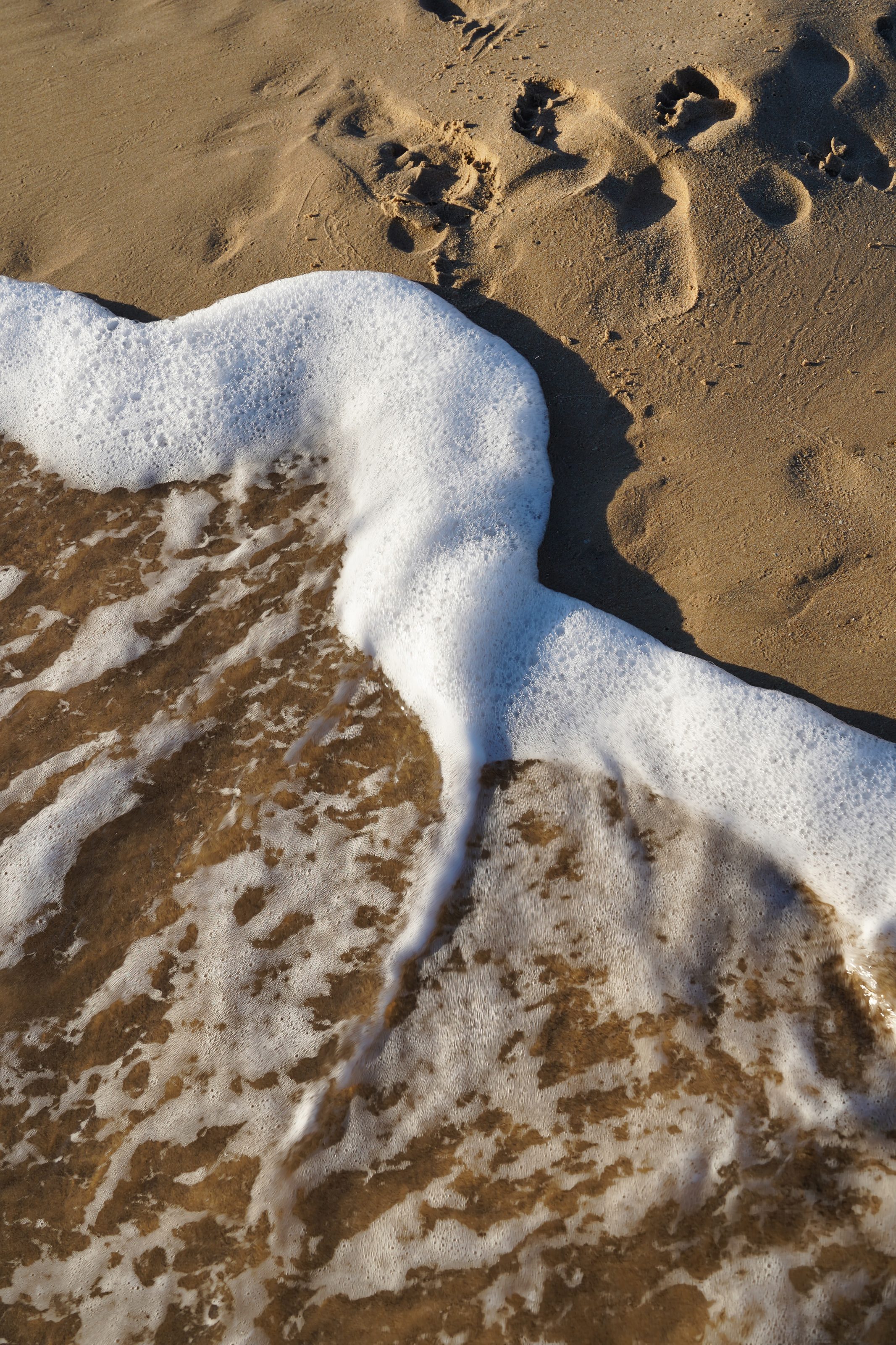 Waves with white foam washing up on a sandy beach, with footprints visible in the sand.