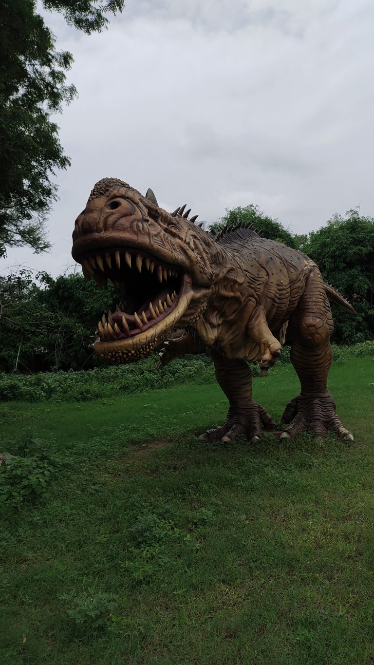 Realistic T-Rex dinosaur model with an open mouth, showcasing sharp teeth, standing in a green park surrounded by trees under a cloudy sky.