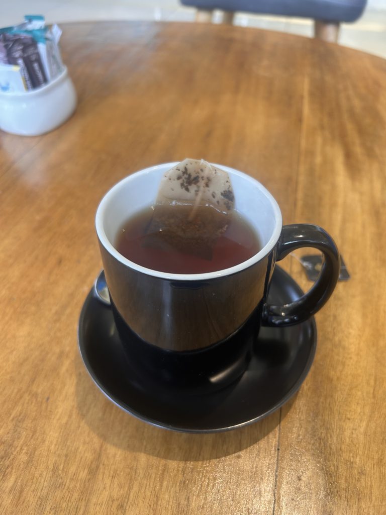 A black cup and saucer with hot tea and a tea bag on a wooden table. In the background, a small container holding sugar packets is visible.