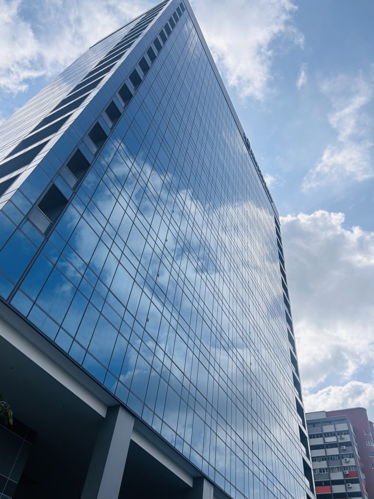 A tall modern hotel with a reflective glass facade, capturing the blue sky and clouds above.