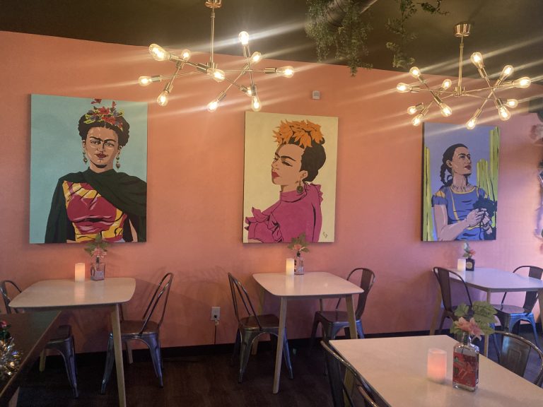 A cozy cafe interior with pink walls features three vibrant paintings. Each painting depicts a woman with colorful attire and accessories. Below the paintings, there are white tables, metal chairs, small floral centerpieces, and dimly lit candles, creating a warm ambiance. Modern chandeliers with multiple exposed bulbs hang from the ceiling.