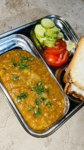 A stainless steel plate containing pav bhaji garnished with cilantro, a side of sliced cucumbers and tomatoes, a lemon wedge, and toasted bread slices.