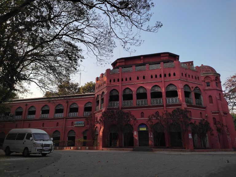 A large, red brick colonial-style building with arched windows and decorative latticework. The structure has multiple levels and features a dome on one side.