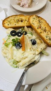A plate of creamy white sauce penne pasta garnished with broccoli, olives, parsley, and a crunchy element, accompanied by slices of garlic bread. 