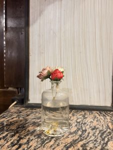 A small bouquet of fabric flowers in a clear glass bottle filled with water, placed on a marbled surface with a light wooden textured background.