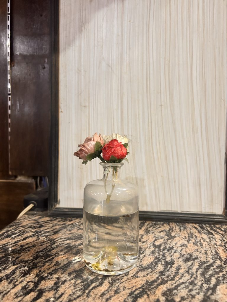 A small bouquet of fabric flowers in a clear glass bottle filled with water, placed on a marbled surface with a light wooden textured background.