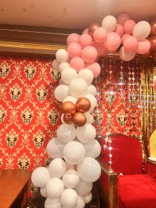 A festive arrangement of white, pink, and metallic bronze balloons forms an arch over a red chair with wooden arms. 