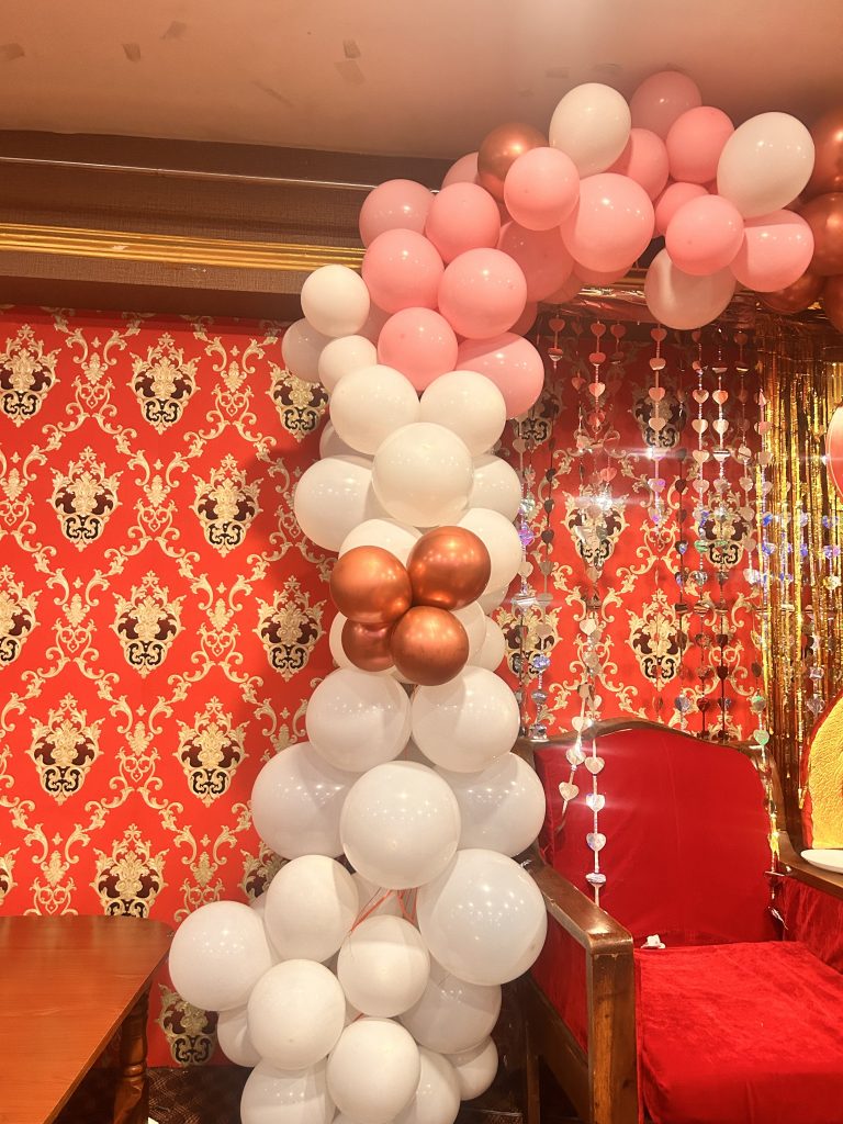 A festive arrangement of white, pink, and metallic bronze balloons forms an arch over a red chair with wooden arms.