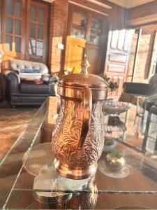 A copper jug with intricate floral engravings sits on a glass table in a warmly lit room. In the background, there is a leather sofa with cushions, a wooden cabinet, and large windows allowing sunlight to stream in.