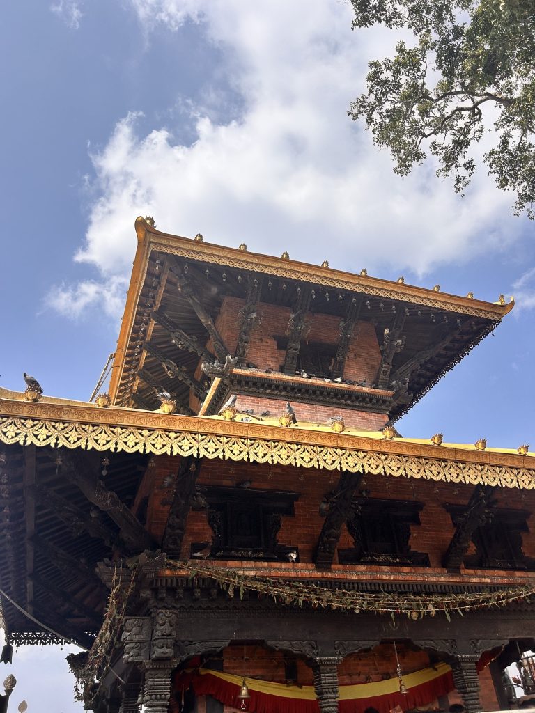 A traditional multi-tiered pagoda-style temple with intricate wooden carvings and golden embellishments, set against a vivid blue sky with scattered clouds