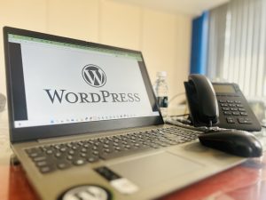 A laptop displaying the WordPress logo on its screen, positioned on an office desk alongside a smartphone, a desk phone, and a bottle of water.