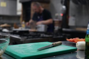 A kitchen knife in the foreground on a cutting board with a cook in the background.