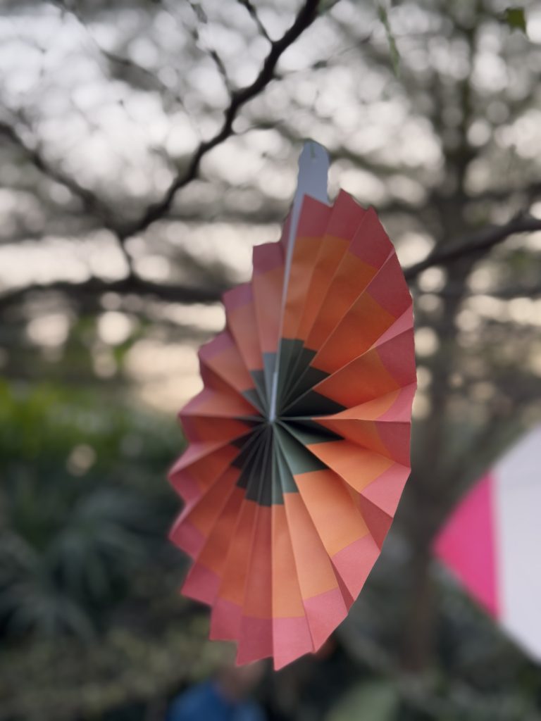 A colorful paper decoration, resembling a folded pinwheel hanging from a tree branch with a blurred background of leaves and branches.