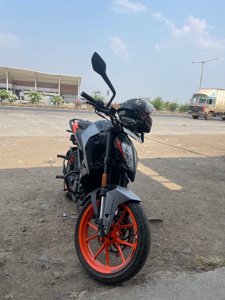 A motorcycle with orange wheels is parked on a dirt surface. A helmet is placed on the handlebar. In the background, there is a road, a building with an awning, and a truck on the street. The sky is clear with some clouds.