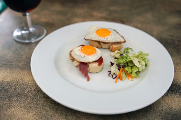 Small toasts with fried quail eggs and Iberico ham with a side of salad. There is a glass of wine in the background.