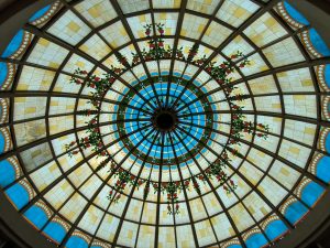 A circular stained glass ceiling with a pattern of geometric shapes, featuring blue and yellow sections. Red and green floral designs radiate from the center.