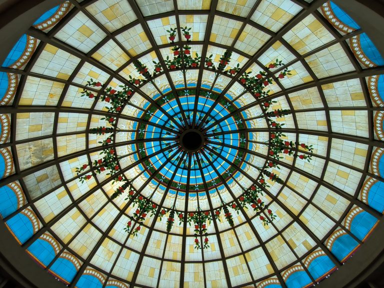 A circular stained glass ceiling with a pattern of geometric shapes, featuring blue and yellow sections. Red and green floral designs radiate from the center.