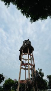 A tall, weathered water tank stands against a cloudy sky, surrounded by lush green trees. 