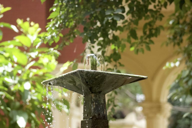 A close-up of a metal fountain with water gently cascading over its edges, surrounded by lush green foliage and a blurred background of architectural arches.