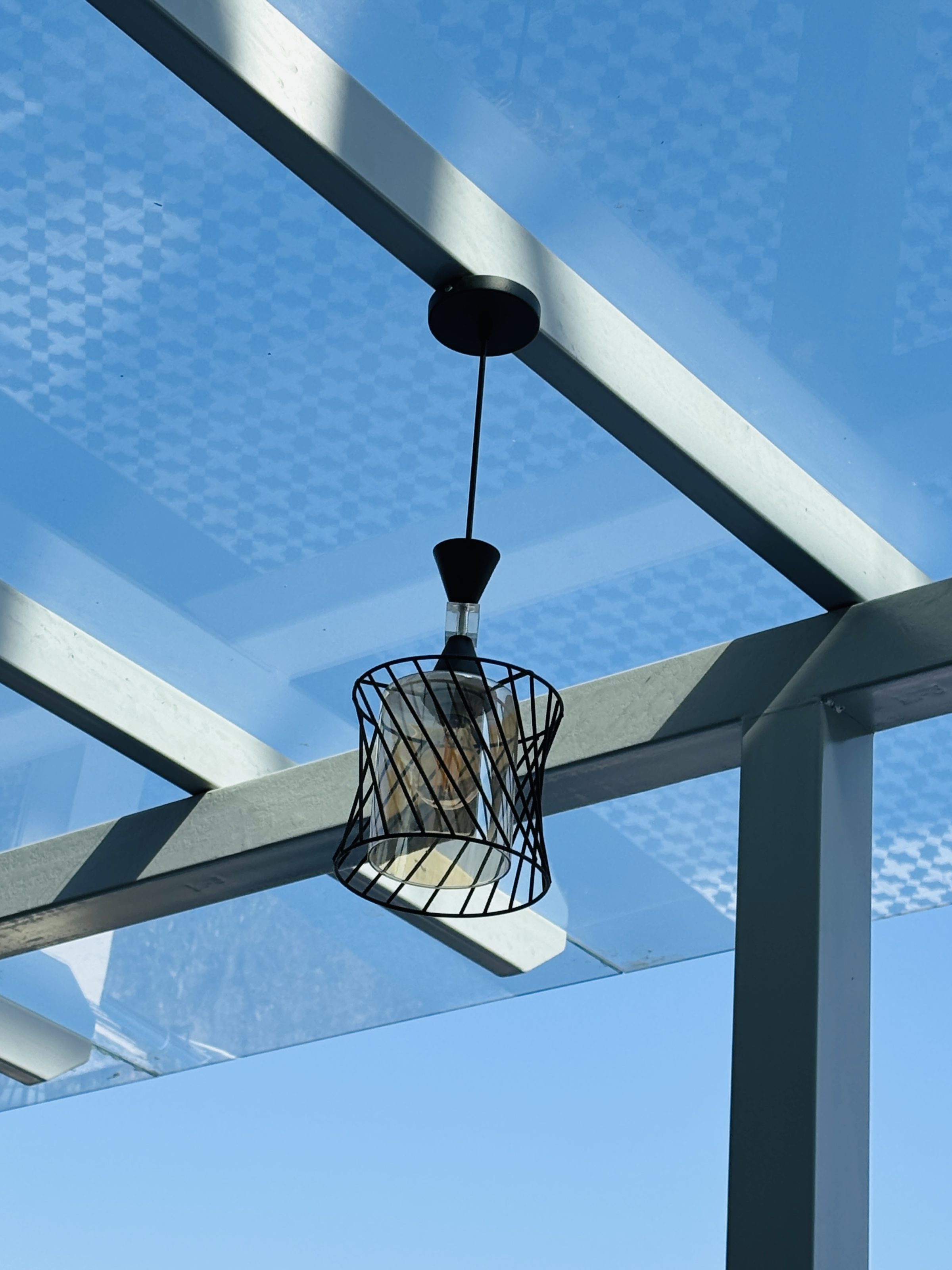 Modern pendant light with a sleek wireframe design hanging under a transparent glass ceiling, reflecting the blue sky and geometric patterns