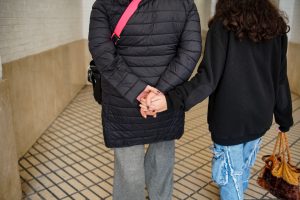 Two individuals walking together, viewed from behind. One person is holding hands with the other person, who is carrying a brown furry bag.