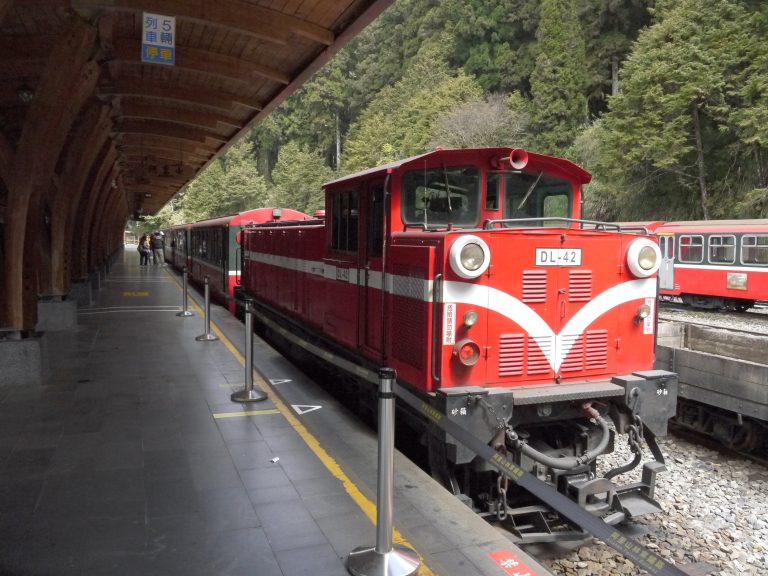 A red train with white accents is stationed on a platform surrounded by forested hills. The platform has a wooden canopy and a few people are walking along it.