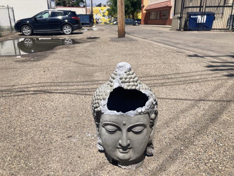 A broken statue of the Buddha’s head exposing the darkness within it sits in the middle of a sunlit alley amid the shadows of wires, a dumpster, and a large puddle.